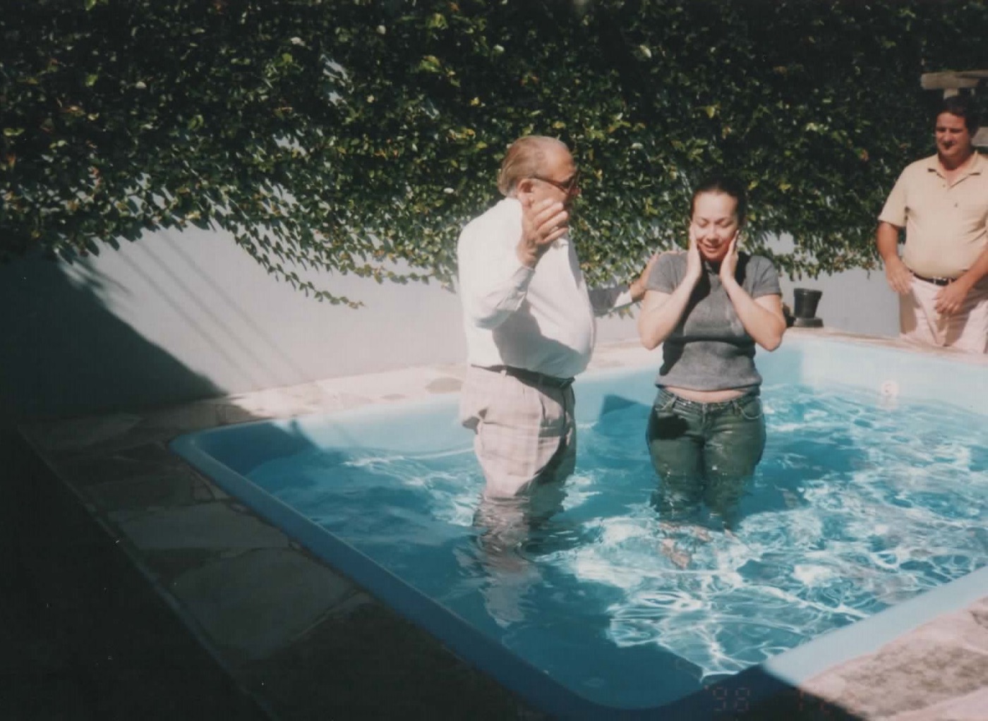 Antenor, de camisa branca, batiza uma jovem em uma piscina em dia de sol, 1998.