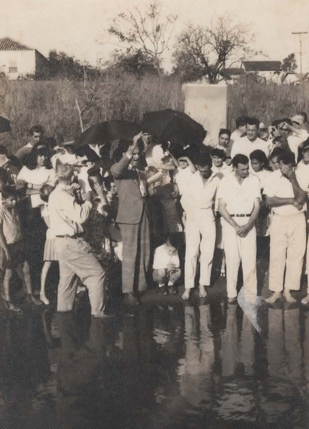 Multidão com guarda-chuvas acompanha da margem um batismo no rio, em foto antiga.