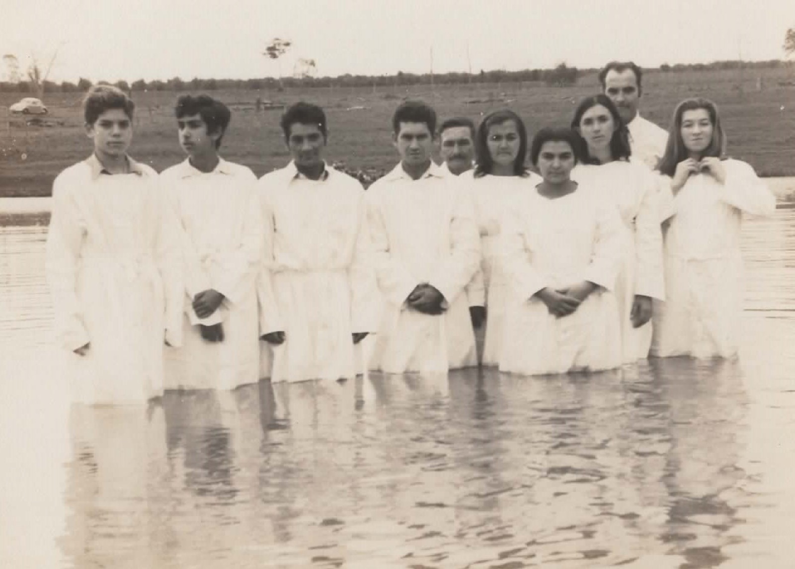 Candidatos ao batismo de túnicas brancas dentro do rio, enfileirados, em foto sépia.