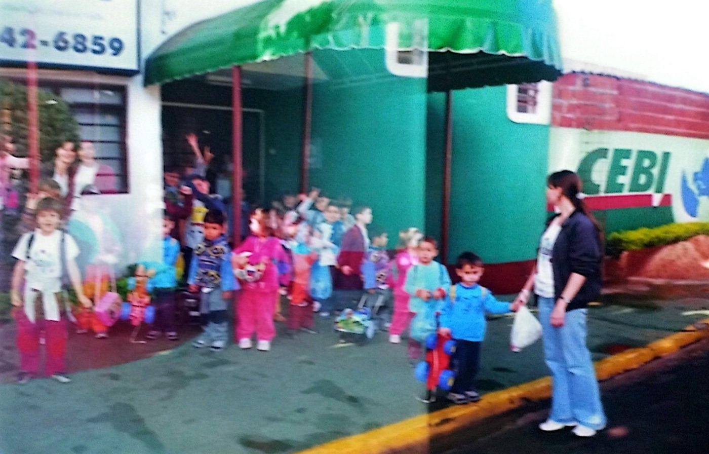 Crianças acenando na porta do CEBI, sob o toldo verde, acompanhadas pelas professoras.