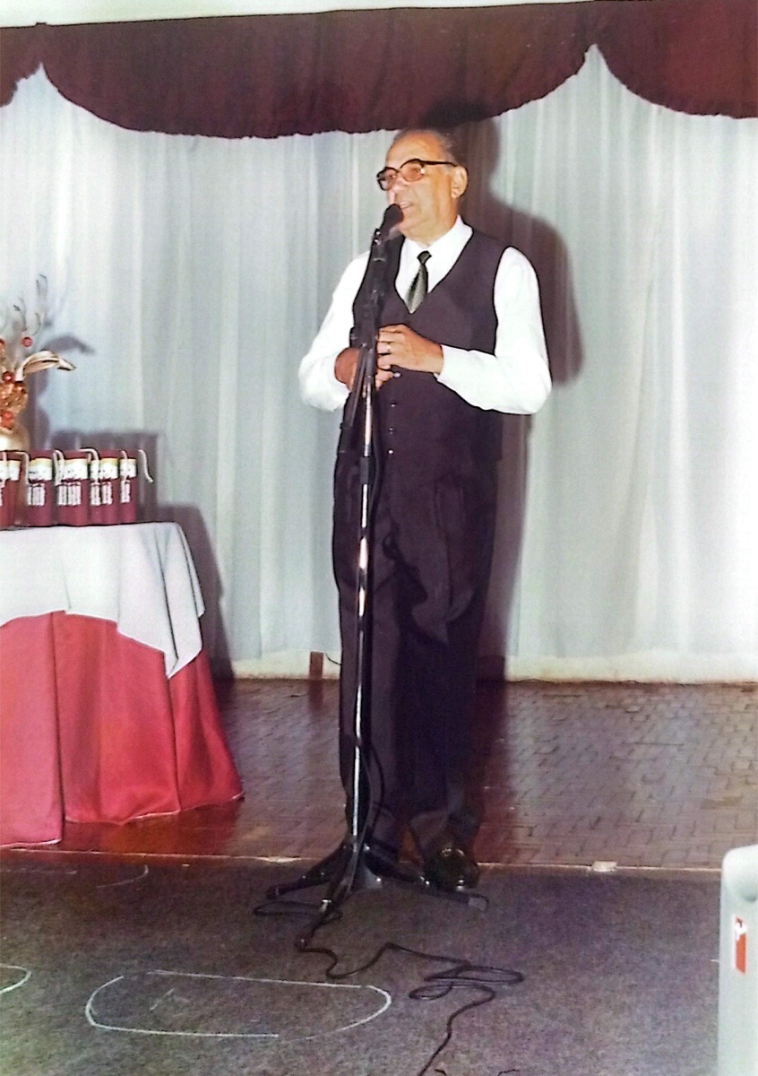 Pr. Antenor discursando ao microfone em uma formatura do colégio, diante de mesa com lembranças dos formandos.