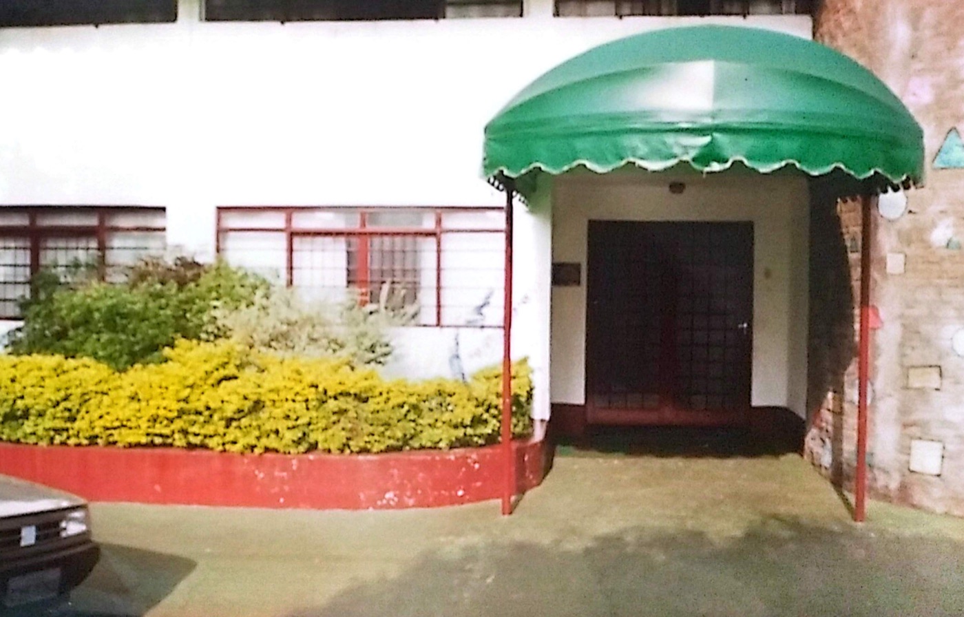 Entrada da primeira sede do CEBI, com toldo verde arredondado e jardim de flores amarelas.