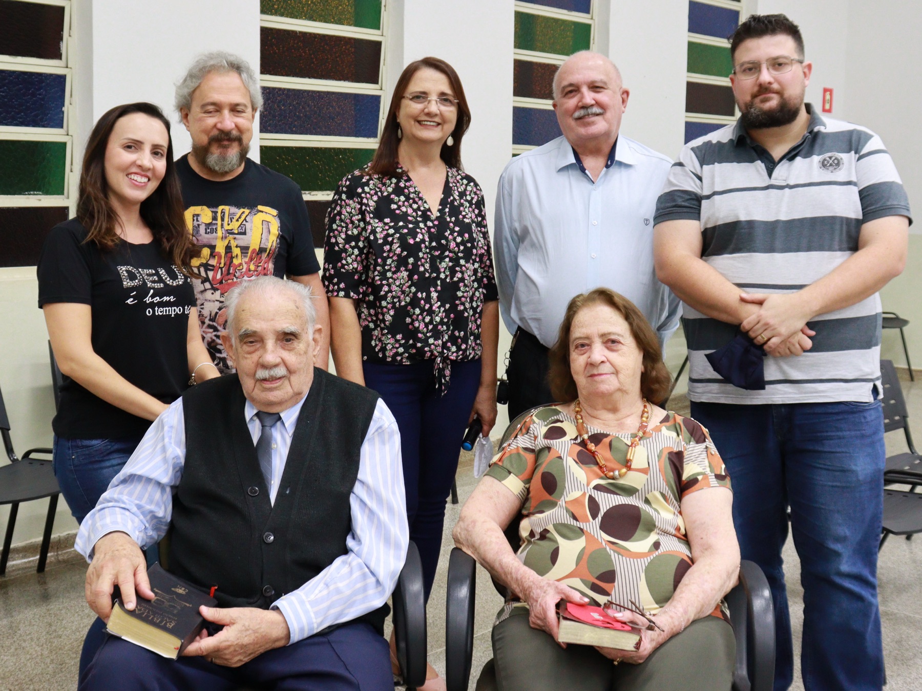 Pr. Antenor e Maria sentados, cercados pela família em pé, diante dos vitrais coloridos da igreja.