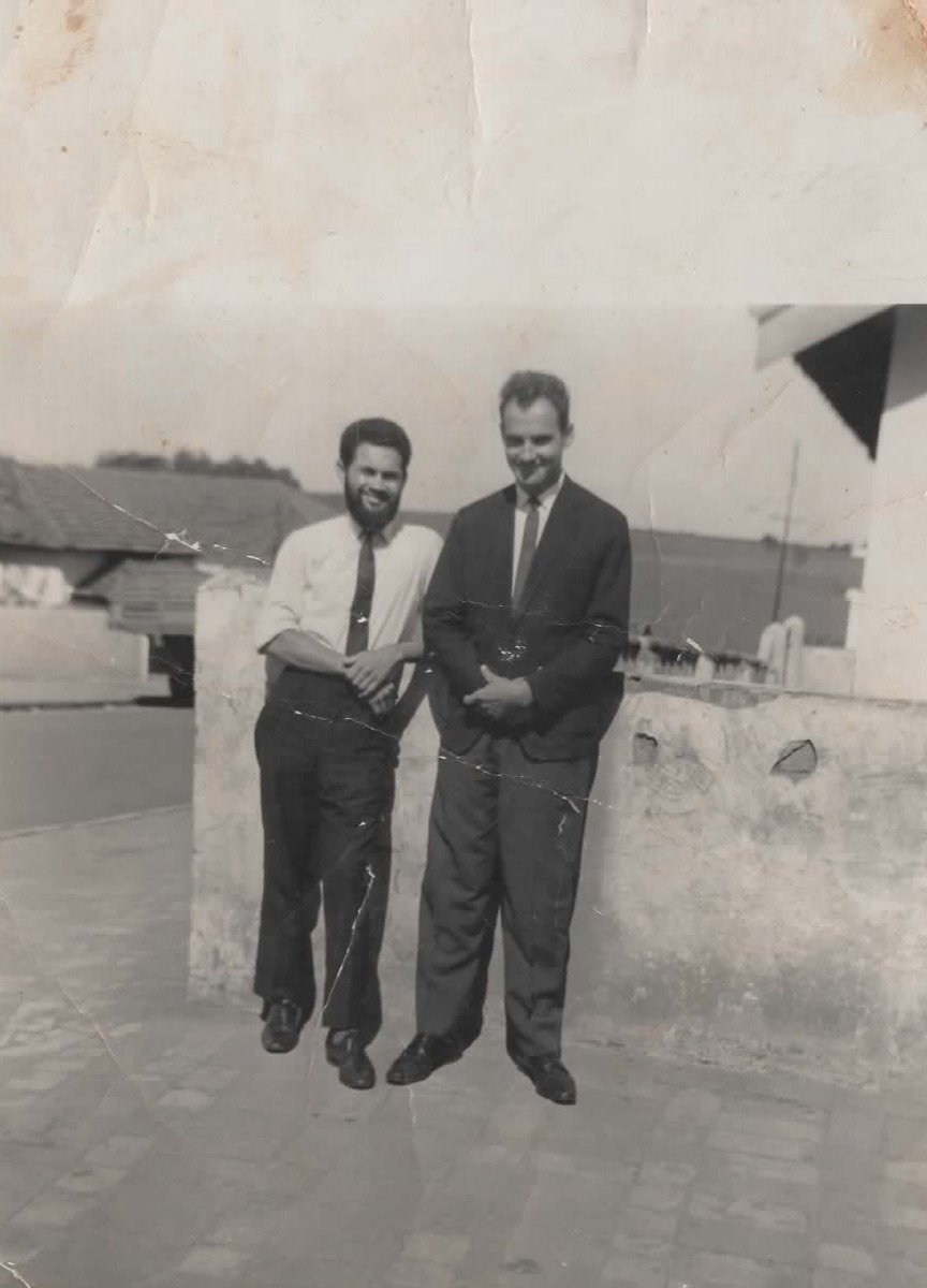 Dois jovens de terno sorrindo sob o sol, em fotografia antiga em preto e branco.