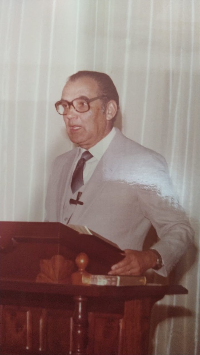 Pastor Antenor pregando ao púlpito, de terno claro, com a Bíblia aberta diante de si.